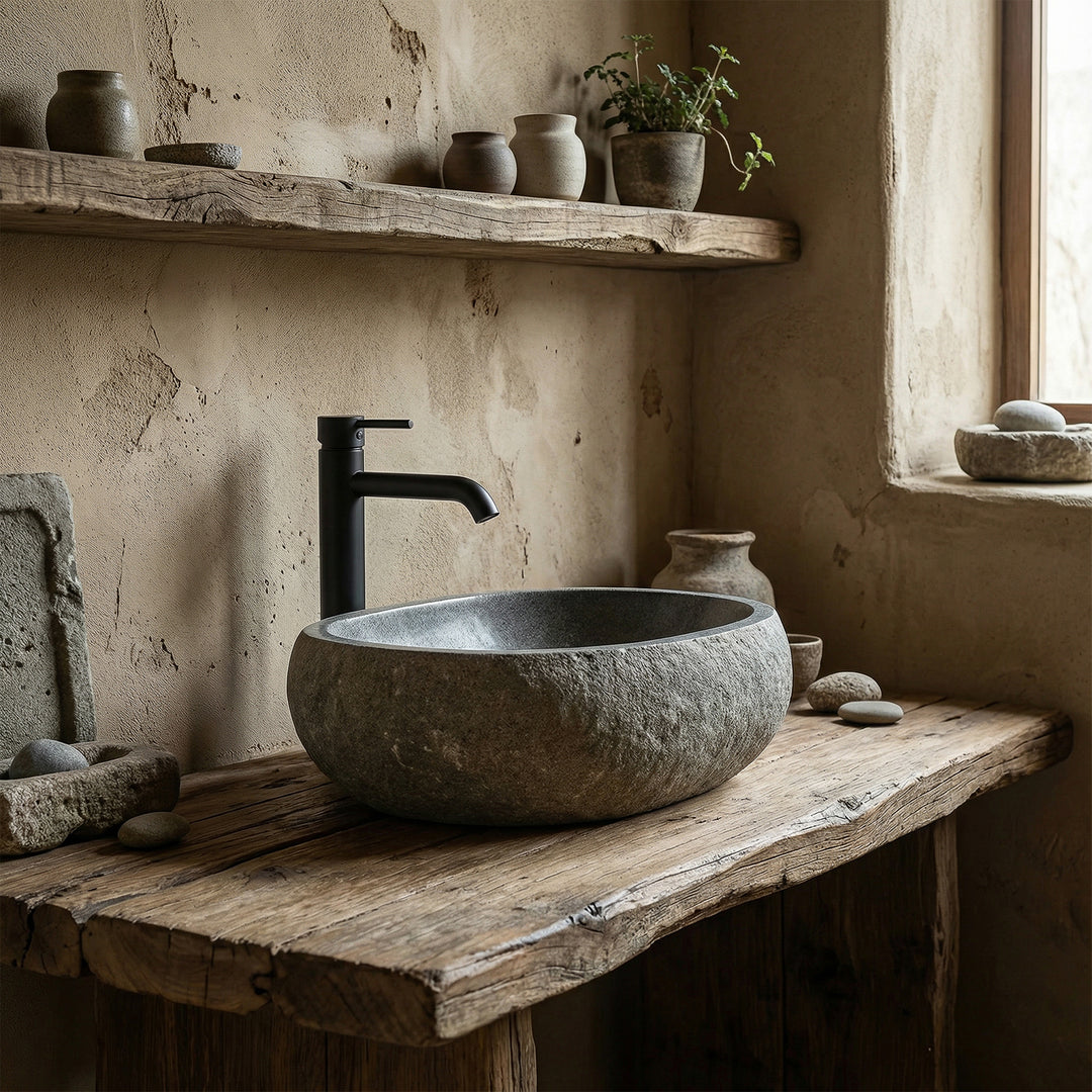 Vasque à poser en pierre de rivière dans une salle de bains naturelle et calme sur une vieille console en bois, associée à des accessoires noirs