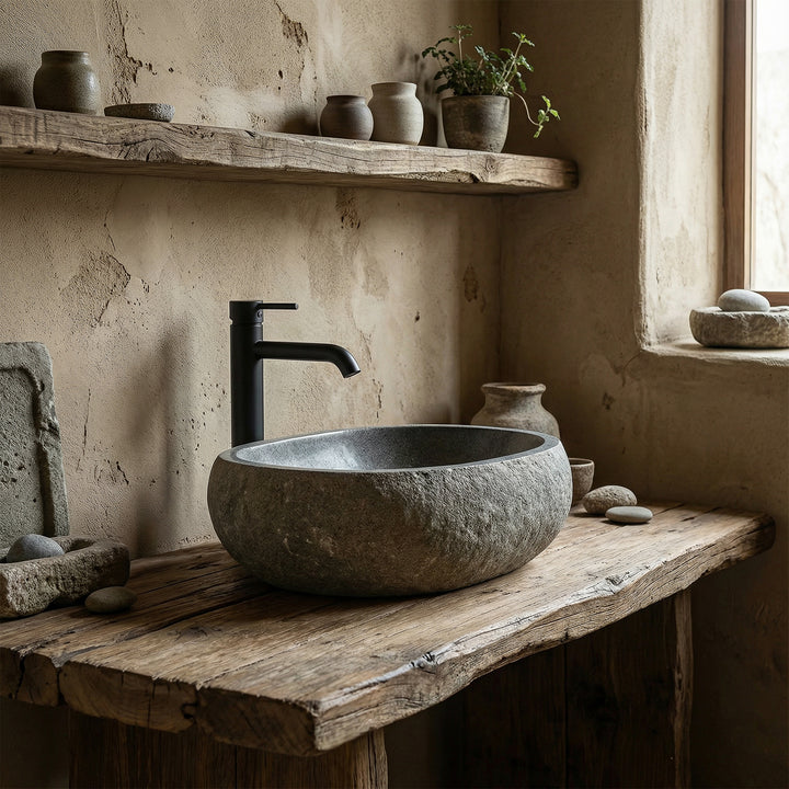 Vasque à poser en pierre de rivière dans une salle de bains naturelle et calme sur une vieille console en bois, associée à des accessoires noirs