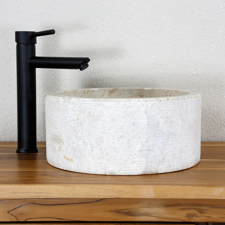Round countertop washbasin made of light natural stone with a fine line structure, combined with a black stand-alone tap.
