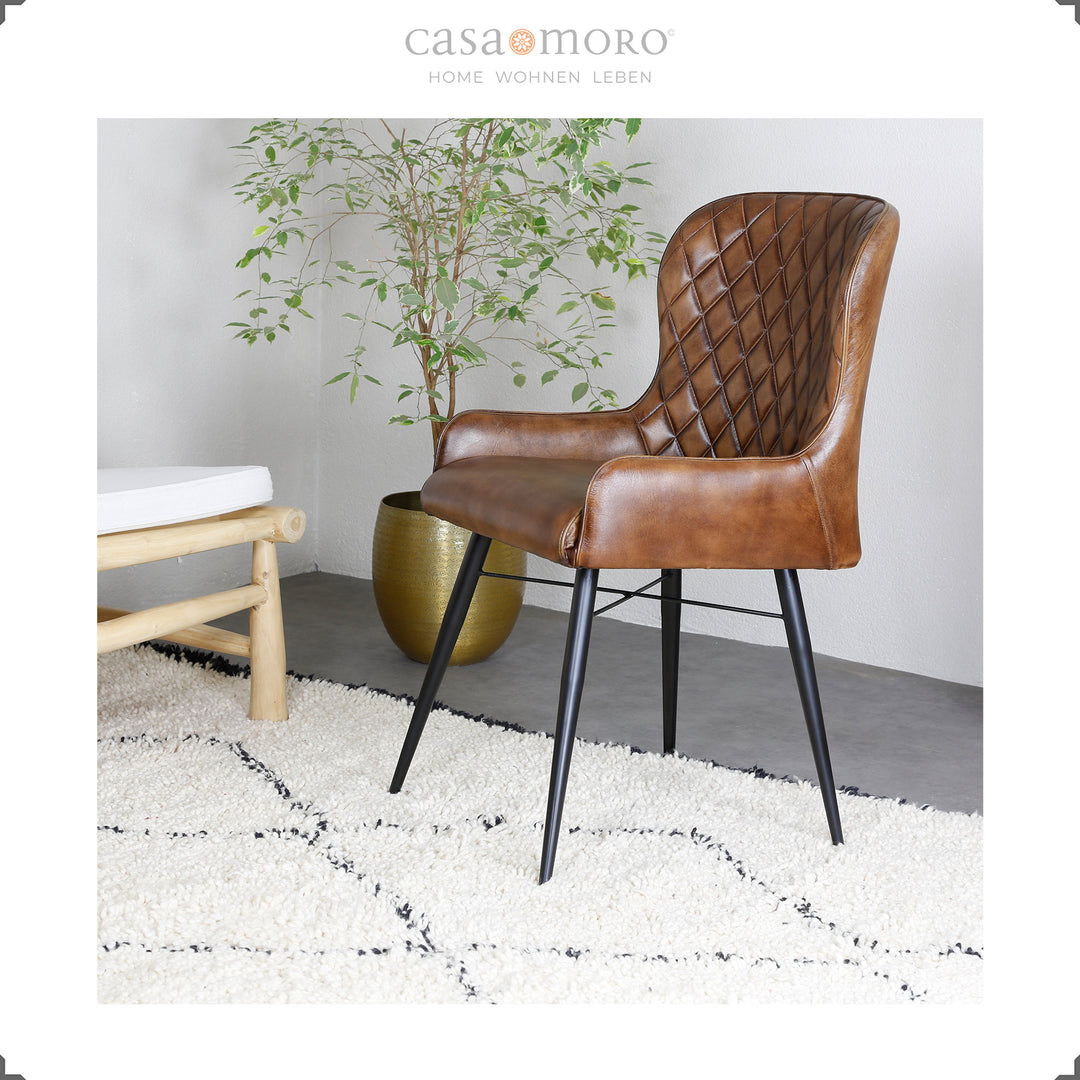 Antique brown leather chair with diamond quilting on a light carpet, next to a golden vase with a green plant - modern living ambience with a natural touch.