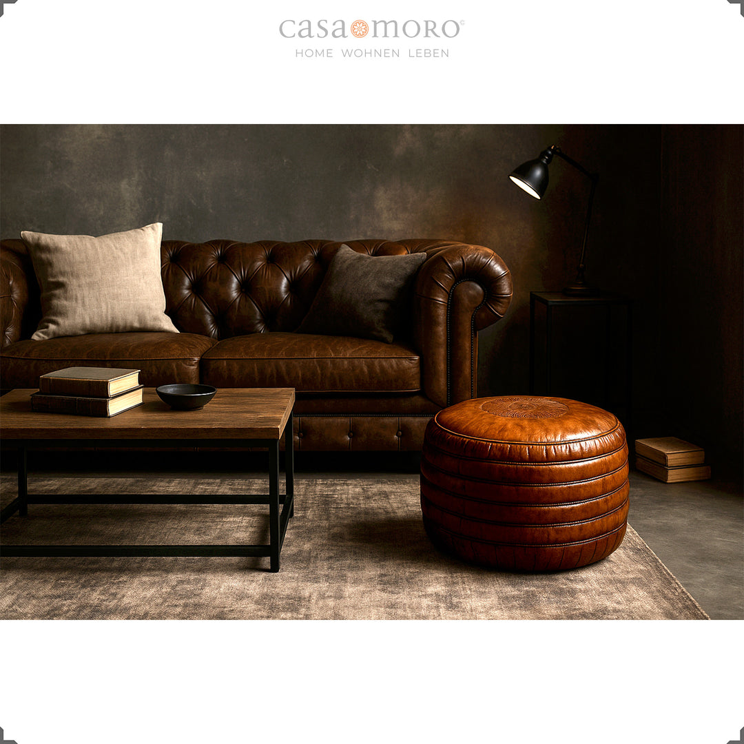 Elegant living room with Chesterfield sofa, coffee table and round leather pouf in brown - stylish scene in industrial and modern-rustic style.