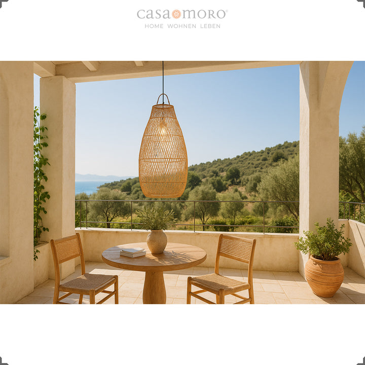 Grande lampe en rotin au dessus d'une table à manger ronde sur une terrasse avec vue sur la verdure et ambiance méditerranéenne