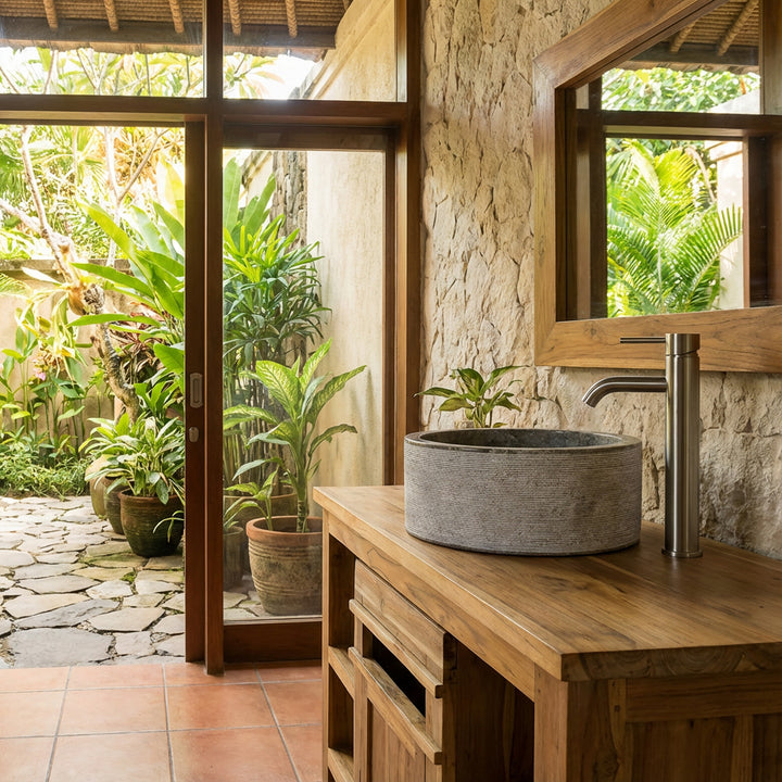 Lavabo rond en pierre grise sur meuble sous vasque en bois, placé dans la salle de bain baignée de lumière avec vue sur le jardin.