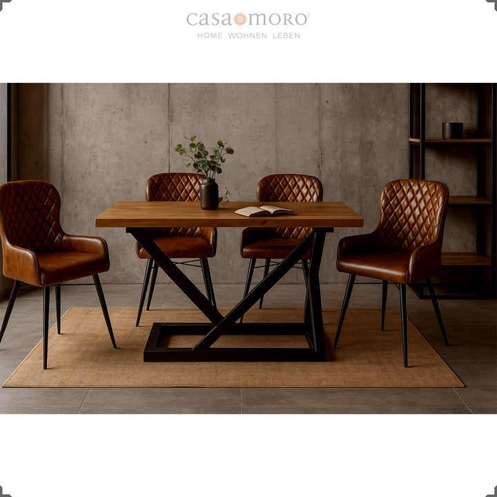 Four antique brown leather chairs with diamond quilting at a modern wooden table with a black metal frame in an elegant loft dining room with a concrete wall.
