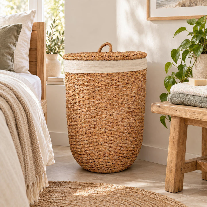 Wäschekorb mit Deckel aus naturfarbenem Seegras und hellem Stoffeinsatz zwischen Bett und Holzbank im Schlafzimmer.